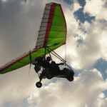 Ultraľahké lietadlo so zelenými krídlami vznášajúce sa na oblohe medzi oblakmi.